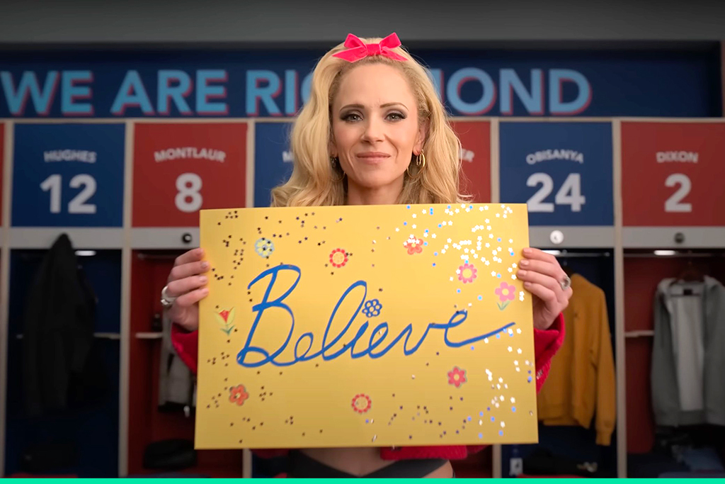 Juno Temple as Keeley Jones holding a glittery sign that says "believe" in the 'Ted Lasso' season 3 trailer.