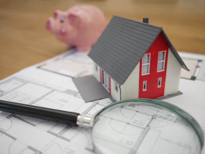 a picture of a magnifying glass on a table with a model of a house, next to building blueprints and a piggy bank. nsw election 